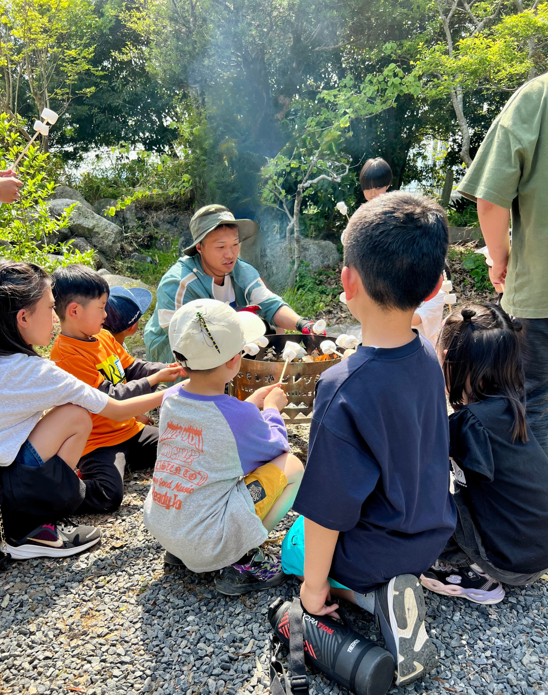MiRAIヒミツ基地デイキャンプで、いっちゃん先生と子どもたちが串に刺したマシュマロを火で炙って楽しんでいる様子。三重県四日市・菰野町エリアで行われた自然体験型の子ども向け習い事イベント。