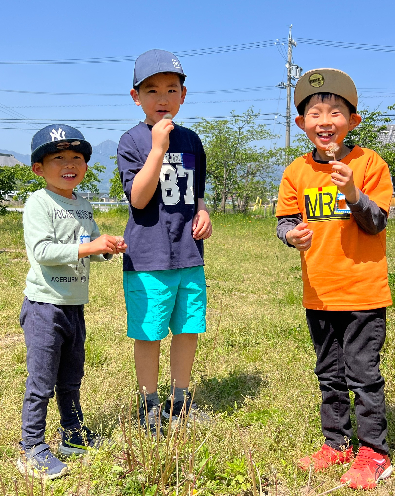MiRAIヒミツ基地デイキャンプで、男の子3人がたんぽぽの綿毛を吹き飛ばしながら笑顔を見せている様子。四日市・菰野町での自然体験型子ども向けイベントの一場面。