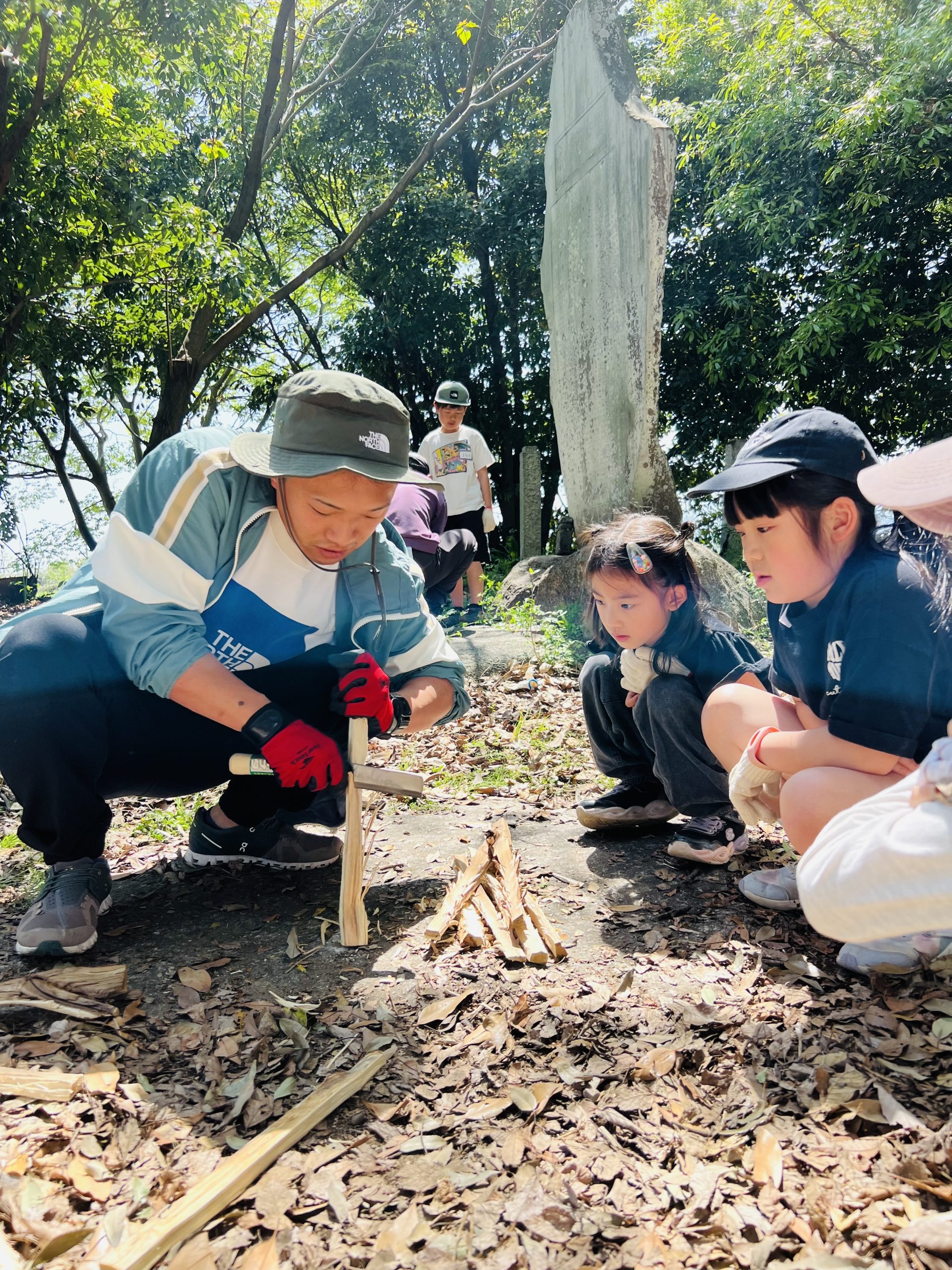 MiRAIヒミツ基地デイキャンプで、講師のいっちゃん先生が子どもたちに「薪に切り込みを入れると火がつきやすくなる」と説明している様子。子どもたちは初めての知識に目を輝かせて真剣に見入っている。