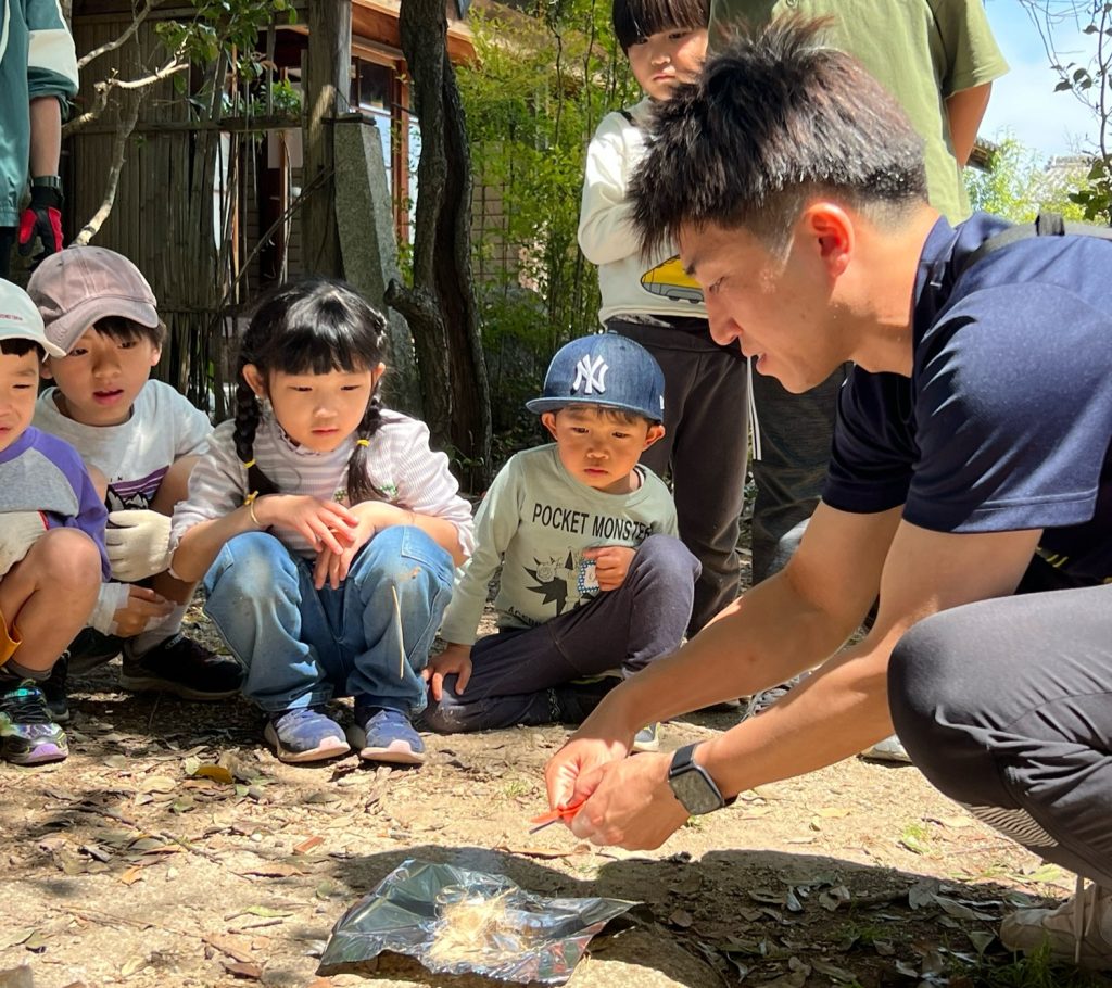 MiRAIヒミツ基地デイキャンプで、けいた先生の火おこしレクチャーを真剣な表情で見つめる子どもたち（三重県菰野町）