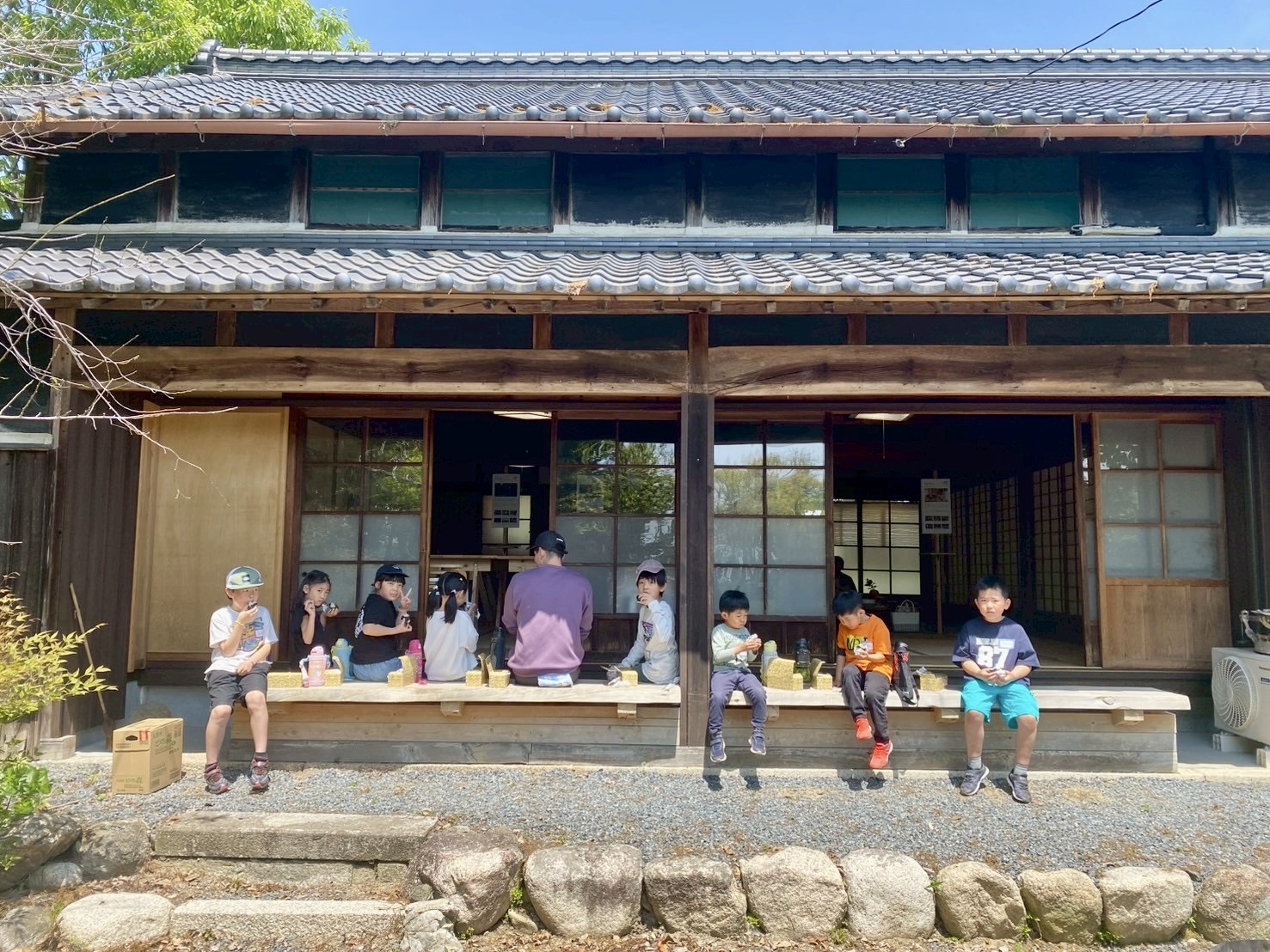 MiRAIヒミツ基地デイキャンプで、子どもたちがかもしかビレッジの古民家の軒下に集まり、お昼ごはんを仲良く食べている様子（三重県菰野町）