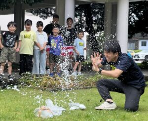 けいた先生がロケットを飛ばす瞬間に水しぶきが上がり、渡り廊下から子どもたちが喜ぶ様子（四日市・小学生・習い事・科学体験）
