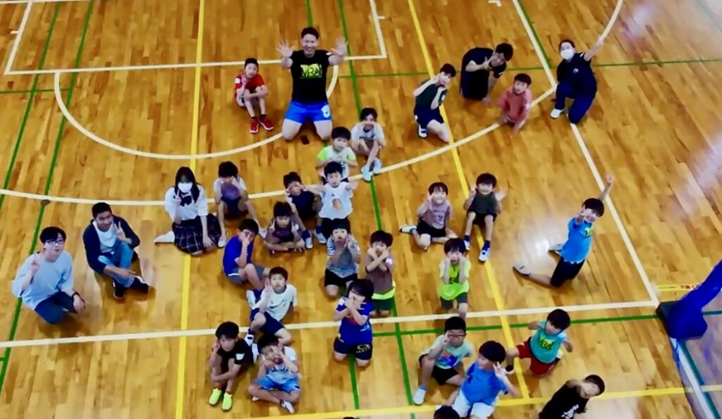 四日市市のメリノール学院で開催されたMiRAIドローン体験イベントの集合写真。体育館でドローンを飛ばして空撮された子どもたちの笑顔