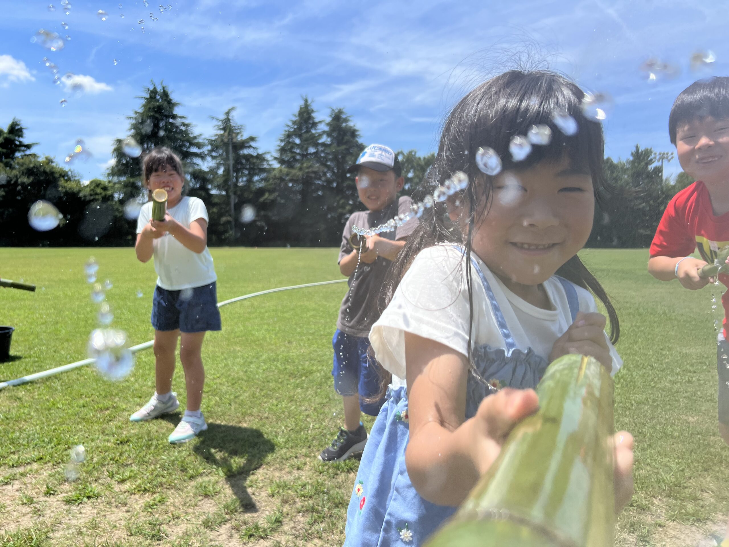 四日市で開催されたMiRAIサマスクで、子どもたちが自作の竹の水鉄砲を使い、天然芝のグラウンドで水を掛け合いながら大笑いして遊んでいる様子。夏休みならではの開放感と笑顔あふれる瞬間。