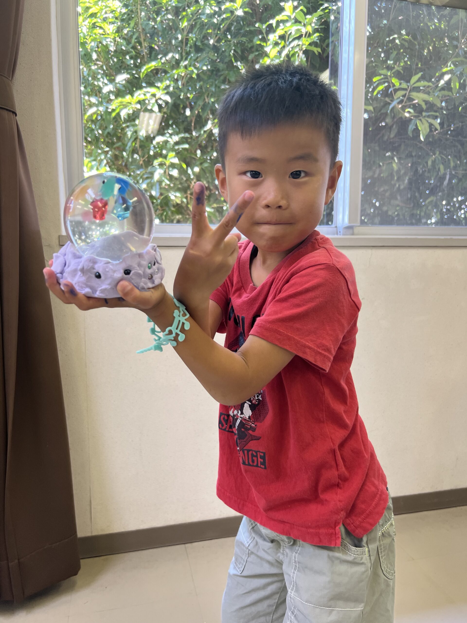 四日市で開催されたMiRAIサマスクで、男の子が自分の手で作ったオリジナルのスノードームを手に持ち、笑顔で記念撮影をしている様子。夏休みの創作体験で達成感と喜びを味わっている瞬間。