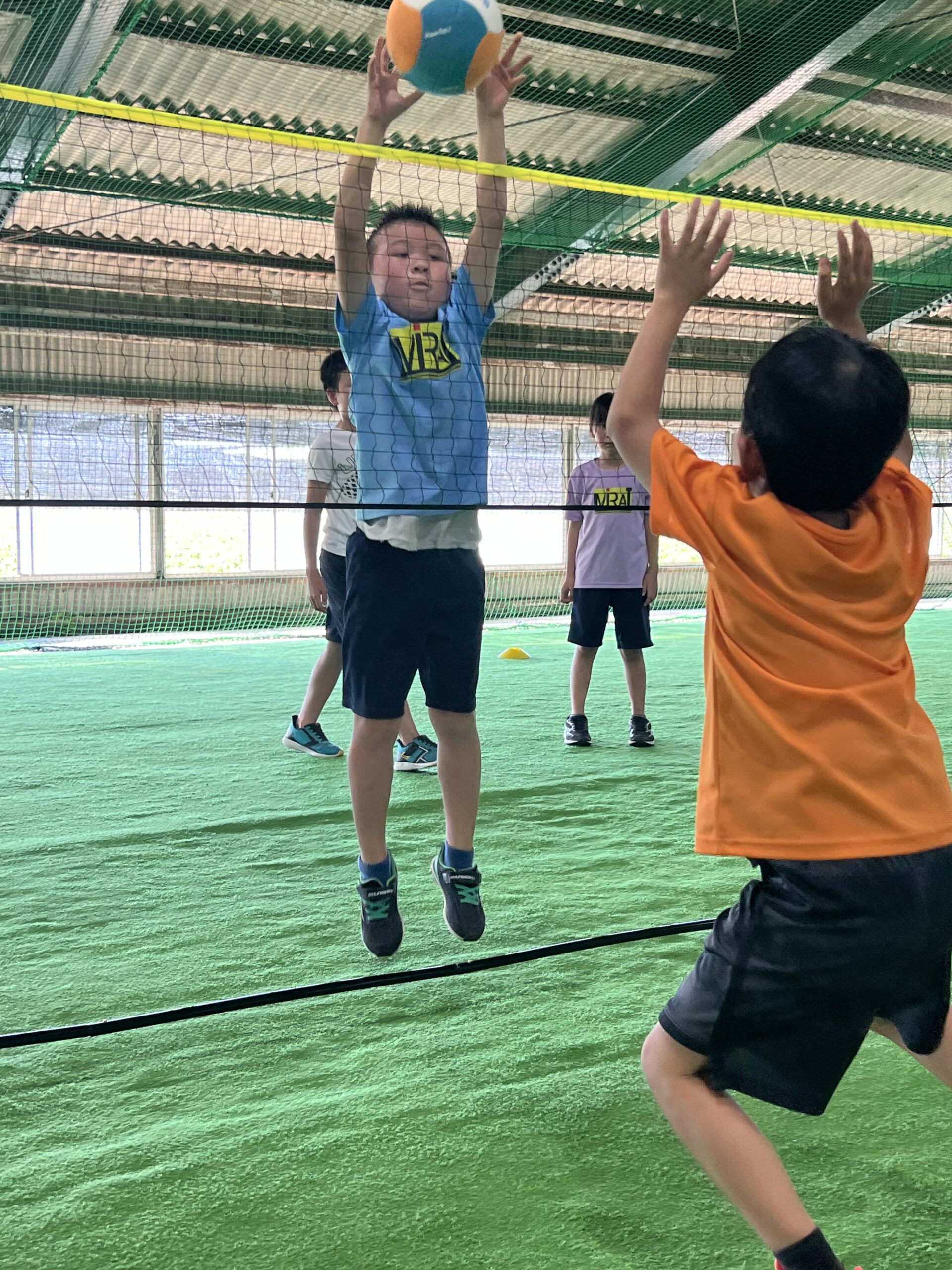 三重県四日市市のスポーツスクールMiRAIで、ネット型スポーツに取り組む小学生の男の子がネットの前で両手を伸ばし、ブロックに挑戦している様子