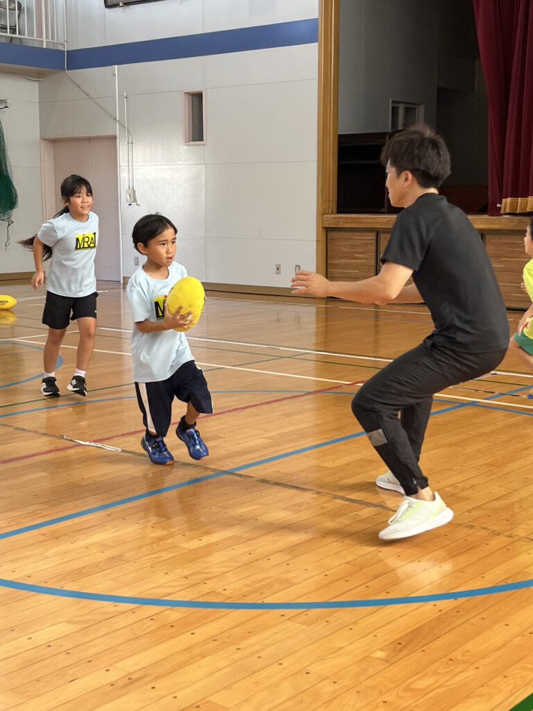 四日市・菰野町エリアの小学生が、総合スポーツスクールMiRAIの体験会でラグビーボールを持ち、真剣な表情でゲームに取り組む様子