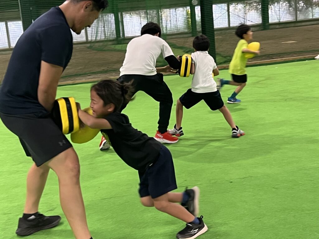 三重県菰野町の総合スポーツスクールMiRAIのラグビー教室で、タックルバッグを持つ先生に子どもが笑顔でタックルする練習の様子