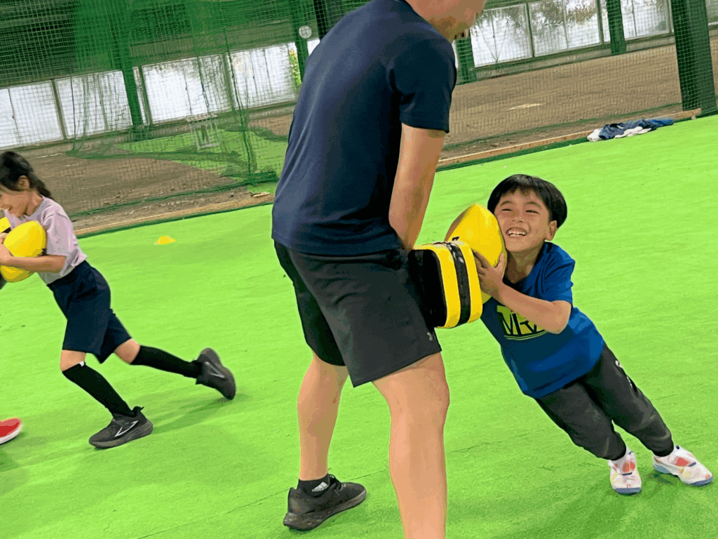 四日市・菰野町エリアの小学生が、総合スポーツスクールMiRAIの体験会でタックルバッグを持つ先生に笑顔でタックルしている様子