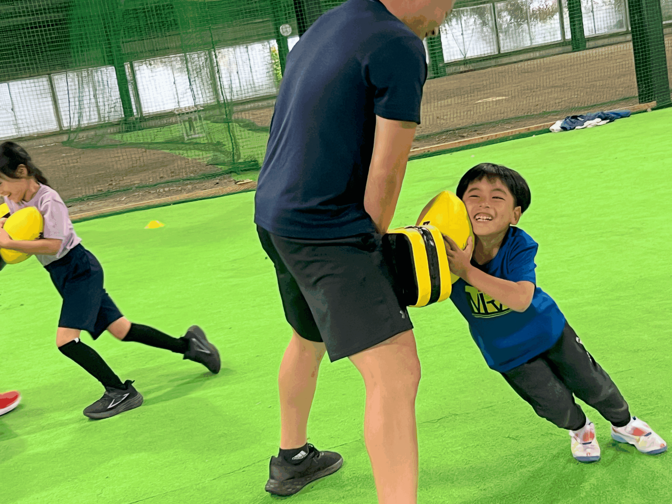 四日市・菰野町エリアの小学生が、総合スポーツスクールMiRAIの体験会でタックルバッグを持つ先生に笑顔でタックルしている様子