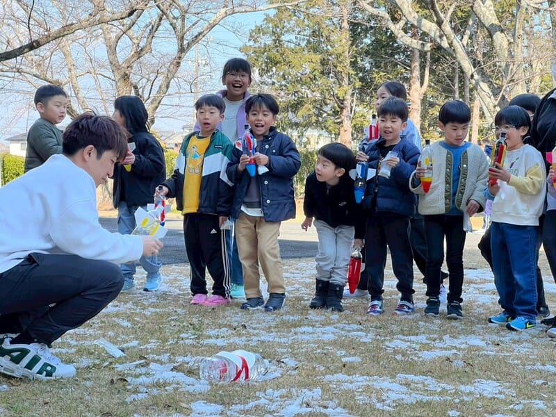 雪の残る校庭で、総合スポーツスクールMiRAIの科学教室の先生がペットボトルロケットの発射準備をお手本として行い、子どもたちが見守っている様子