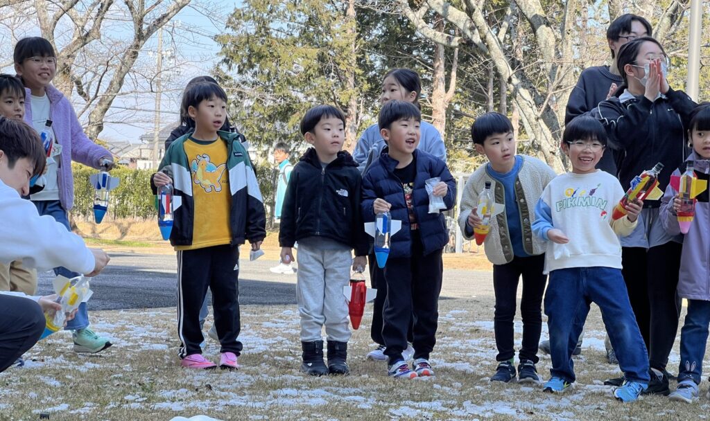 雪の残る校庭で、総合スポーツスクールMiRAIの科学教室の先生がペットボトルロケットの発射準備をお手本として行い、子どもたちが見守っている様子