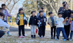 雪の残る校庭で、総合スポーツスクールMiRAIの科学教室の先生がペットボトルロケットの発射準備をお手本として行い、子どもたちが見守っている様子